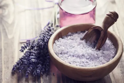 Lavendel und Kristallsalz in einer Holzschale, perfekt zum **Badesalz selber machen**. Ein Glas mit lila Flüssigkeit steht daneben.