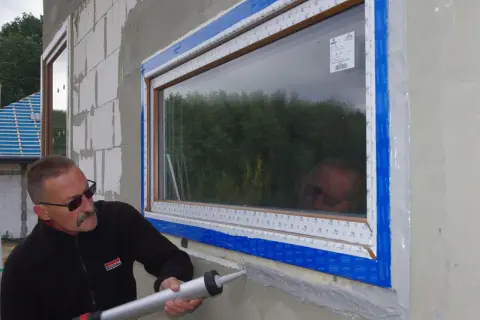Fachowiec z logo Soudal zapewnia szczelny montaż okien, aplikując masę uszczelniającą.