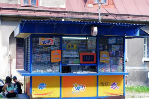 Kiosk Lotto z ekspozycją zdrapek i informacją o kumulacji. Dzieci siedzą obok, zastanawiając się, dlaczego kolektorzy nie chcą sprzedawać zdrapek z konkretnych numerów paczek.