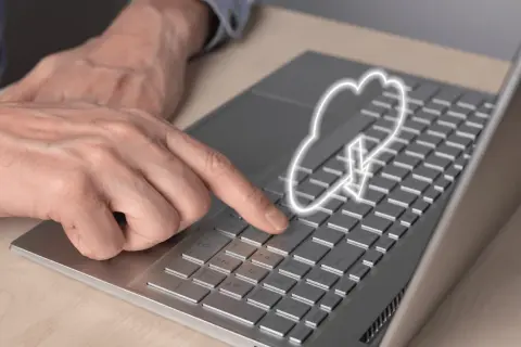 Dłoń naciska klawisz na laptopie, nad którym unosi się symbol chmury internetowej z ikoną pobierania.