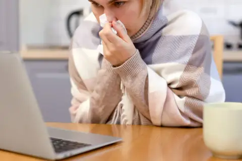 Chora kobieta z chusteczką przy nosie, owinięta kocem, sprawdza w internecie, ile wynosi zasiłek chorobowy.