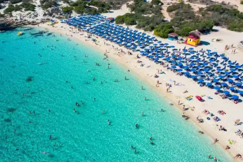 Słoneczny dzień na plaży Makronissos. Turkusowa woda, biały piasek i mnóstwo niebieskich parasoli. Ludzie kąpią się i opalają.