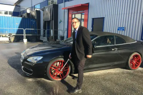 Man i kostym med glasögon vid en svart cabriolet med röda fälgar. Han håller i en fälgnyckel, redo att dra åt efterdra hjulbultar.