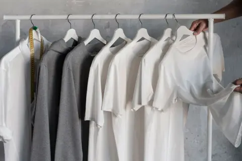 En vit skjorta, grå hoodie och flera vita t-shirts hänger på en klädställning. En hand plockar ner en av t-shirtarna, som en del av en basgarderob herr.