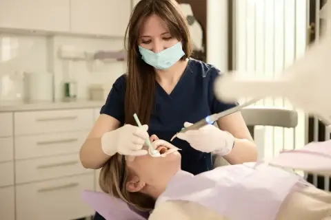 Dentystka w masce i rękawiczkach wykonuje zabieg stomatologiczny u pacjentki.