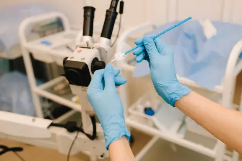 Ręce w niebieskich rękawiczkach przygotowują próbkę do badania ginekologicznego pod mikroskopem.