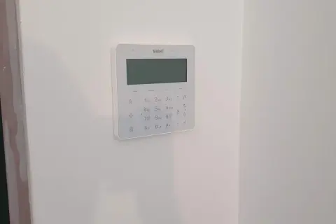 Biały panel alarmowy z klawiaturą numeryczną i ekranem na białej ścianie.