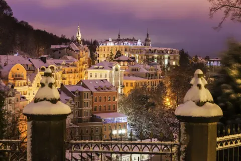 Winterliche Pracht in Karlsbad: verschneite Gebäude unter violettem Abendhimmel, beleuchtet von warmem Licht.
