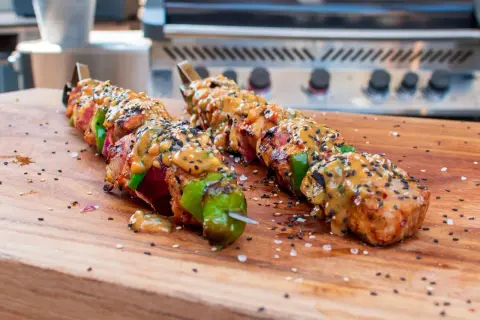 Fertige Schaschlik-Spieße mit Sauce und Sesam auf einem Holzbrett, bereit für den Schaschlik Grill.