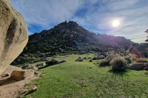 Majestuosa montaña rocosa en la ruta Soto del Real, con un prado verde y el sol brillando en un cielo azul con nubes.