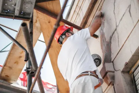 Obrero con casco rojo repara grietas en una fachada desde andamios.