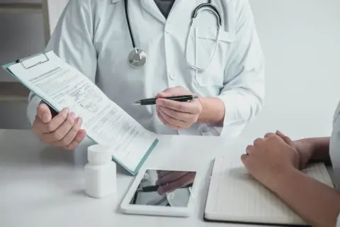 Lekarz w białym kitlu omawia z pacjentem dokumenty, prawdopodobnie dotyczące zwolnienia lekarskiego w Norwegii. Na stole leżą tablet i notatnik.