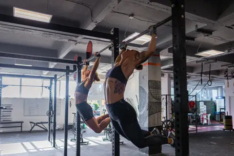 Dwie kobiety wykonują podciąganie na drążku w siłowni. To część treningu, który może być elementem "fran crossfit co to jest".
