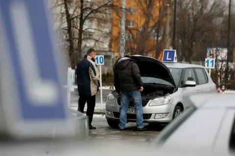 Dwóch mężczyzn przy samochodzie z otwartą maską, znaki "L" i numery na placu manewrowym.