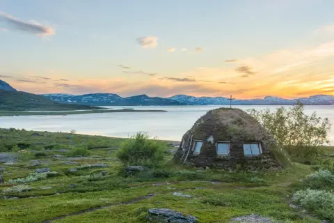 En grästorvstuga med kors på toppen vid en sjö i Padjelanta nationalpark, Sverige, under en solnedgång.