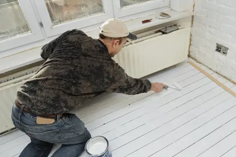 Un homme peint un plancher en bois blanc. Il utilise un rouleau et un pot de peinture. La question "peut on peindre du stratifié" est pertinente pour ce type de projet.