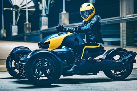 Kierowca w żółtym kasku na futurystycznym motorze trójkołowym Can-Am, gotowy do jazdy.
