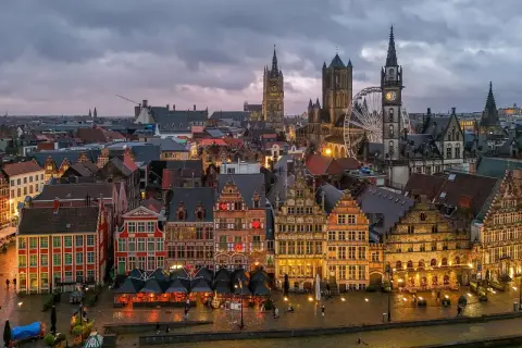 Gryning över Gent, Belgien. Historiska byggnader längs en kanal, med ett pariserhjul och klocktorn som reser sig mot en molnig himmel.