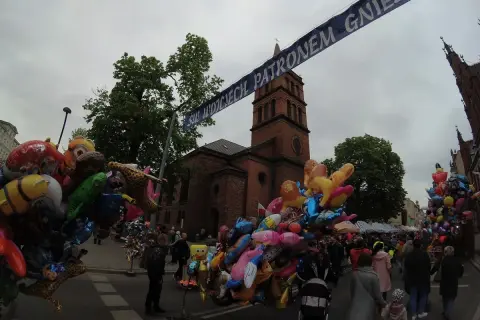 Kolorowe balony z postaciami z bajek i zwierzątkami, idealne gniezno atrakcje dla dzieci, na tle ceglanej wieży kościelnej i tłumu ludzi.