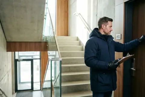 Mężczyzna w zimowej kurtce puka do drzwi mieszkania z tabliczką "402", trzymając teczkę.