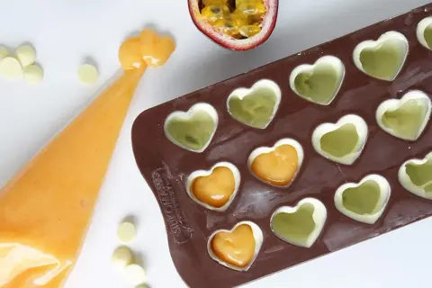 Gör egna enkla chokladpraliner recept med passionsfrukt och vit choklad i hjärtformad form. En påse med orange fyllning ligger bredvid.
