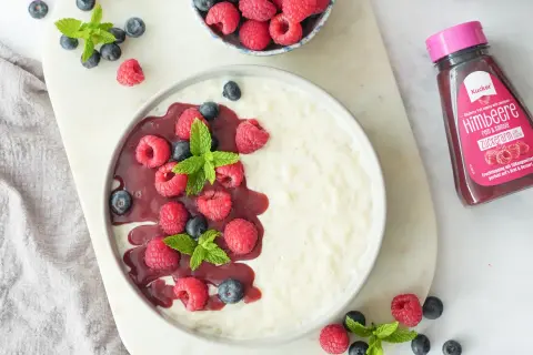 Cremiger Milchreis ohne Zucker, garniert mit frischen Himbeeren, Blaubeeren und Minze. Dazu eine Flasche zuckerarmer Himbeer-Fruchttoppping.