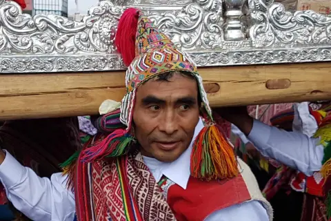 Kolorowy strój i tradycyjna czapka zdobią jednego z mężczyzn niosących ozdobną ramę. To **Indianie z Cuzco** podczas uroczystości.