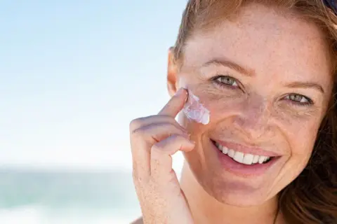 Frau mit Sommersprossen trägt Sonnenfluid auf ihr Gesicht auf, um ihre Haut vor der Sonne zu schützen.