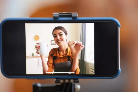 Frau hält Tasse und Keks, während sie ihr **iPhone als Webcam** für ein Kochvideo nutzt.