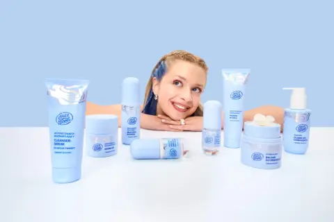 Uśmiechnięta dziewczyna z niebieskimi pasemkami otoczona kosmetykami Face Boom. Wśród nich kremy i sera, które dzięki caprylyl glycol odświeżają skórę.