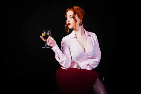 Donna con capelli rossi tiene un calice di vino da meditazione, con un'espressione pensierosa. Indossa una camicetta rosa e una gonna bordeaux.