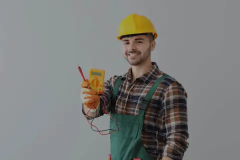 Uśmiechnięty elektryk w żółtym kasku i ogrodniczkach trzyma miernik. Dowiedz się, ile kosztuje przegląd instalacji elektrycznej.