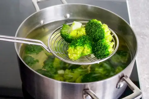Frisch geernteter, wilder Brokkoli wird in einem Topf mit kochendem Wasser blanchiert. Ein Sieb hebt die leuchtend grünen Röschen hervor.