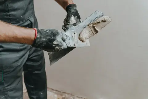 Ręce w rękawiczkach nakładają gładzie na ścianę za pomocą szpachli.