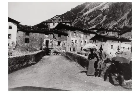 Un villaggio di montagna immortalato da Giovanni Verga fotografo. Coppie eleganti passeggiano con ombrelli, sullo sfondo case in pietra e pendii scoscesi.