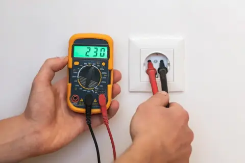 Un multimètre affiche 230V, tandis que ses sondes sont insérées dans une prise. La vérification de la prise terre est obligatoire pour la sécurité.