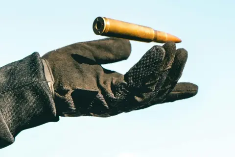 Una mano con guantes tácticos sostiene un cartucho dorado contra un cielo azul claro.