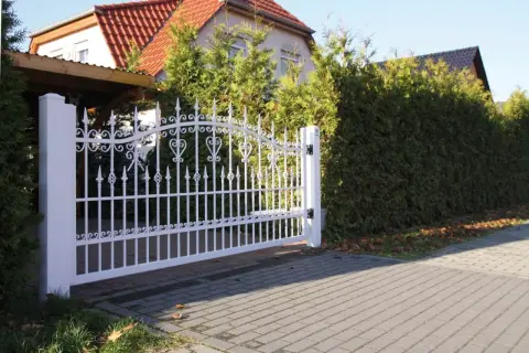 Biała, ozdobna brama wjazdowa, niczym wiejski płot, otwiera drogę do domu z czerwonym dachem, otoczonego zielenią.
