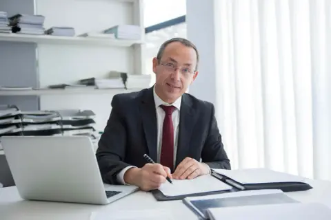 Mężczyzna w garniturze pisze coś na papierze, obok laptop i stosy dokumentów. Jego zaangażowanie w pracę jest widoczne.