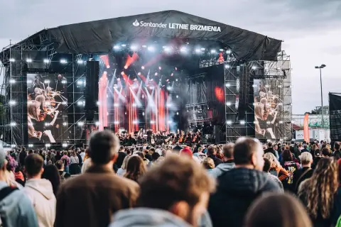 Koncert Santander Letnie Brzmienia z orkiestrą na żywo i ekranami pokazującymi muzyków. Tłum widzów czeka na występ.