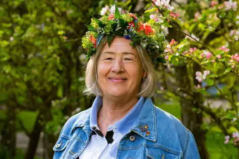 Glad kvinna med blomsterkrans, Bettan byvald, ler i solsken bland blommande träd.
