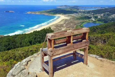 Banco de madera en un monte ventoso con vistas a una playa, el mar azul y un lago.