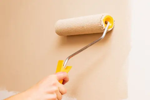 Mano che usa un rullo per pitturare una parete di color sabbia. Impara come pitturare con il rullo per un risultato perfetto.