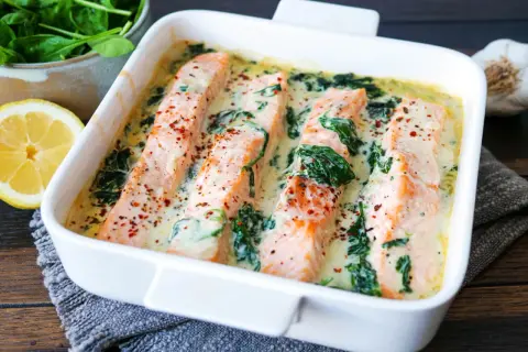 Vier saftige Lachsfilets in cremiger Spinatsauce, bestreut mit Gewürzen, in einer weißen Auflaufform. Daneben liegen frischer Spinat und eine Zitronenspalte.
