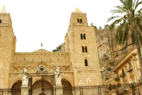 La imponente catedral de Cefalú se alza majestuosa contra un acantilado rocoso, con una palmera que añade un toque tropical a la escena.