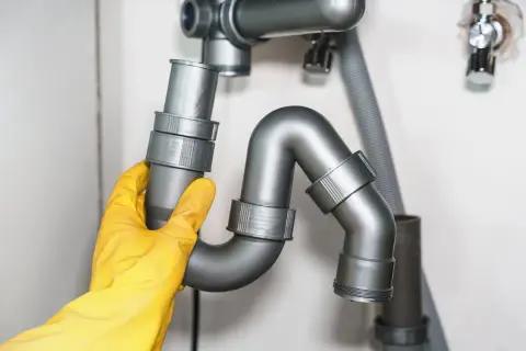 Main d'un plombier en gant jaune ajustant un siphon de rejet d'eau PVC sous un évier.