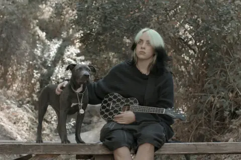 Billie Eilish z pieskiem i ukulele w rękach, siedząca na ławce w otoczeniu natury.
