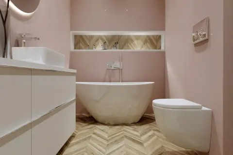 Elegancka łazienka z wolnostojącą wanną i baterią wannową. Jasnoróżowe ściany, drewniana podłoga w jodełkę.