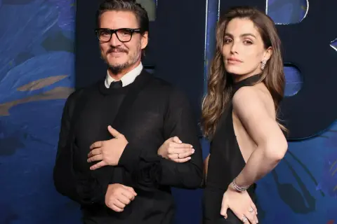 Pedro Pascal y una mujer elegante posan juntos. El actor, con gafas y un suéter negro, luce un cabestrillo. Ella, con un vestido negro y joyas brillantes, sonríe.