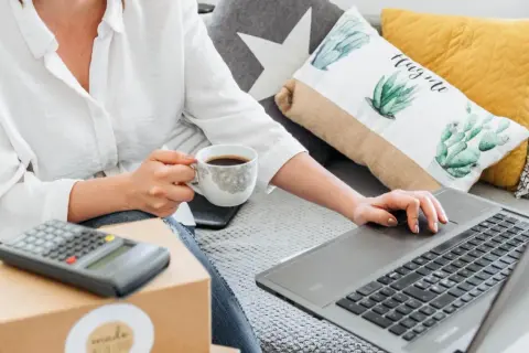 Kobieta z kawą uczy się, jak sprzedawać na OLX, korzystając z laptopa i kalkulatora.